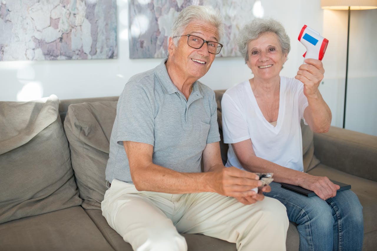 Happy senior couple sitting on a couch, sharing a joyful moment while holding health-related devices in a well-lit living room.